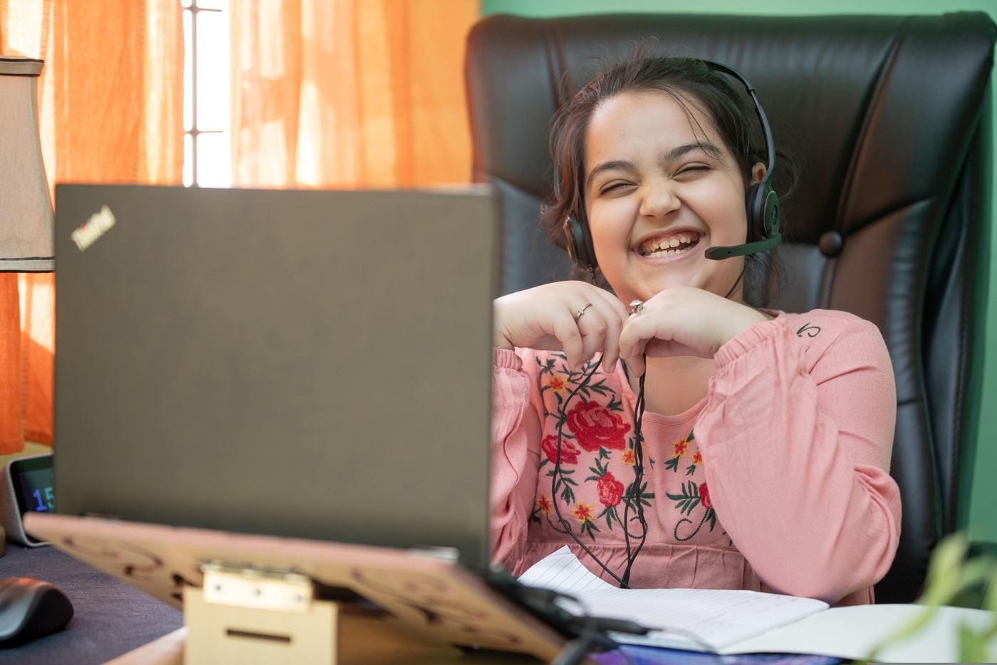 A girl laughing during a live online lesson, wearing a headset at her laptop